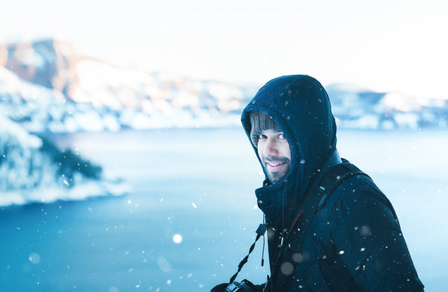 man in hooded snow jacket smiling overlooking water and mountains in the snow