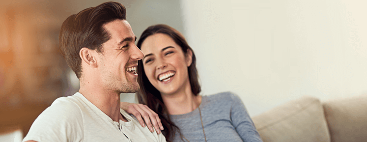 brunette man and brunette woman sitting on beige couch and laughing