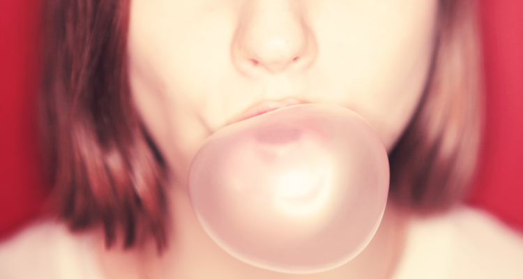 Close-up of the mouth of a brown-haired woman blowing a pink gum bubble against a red wall