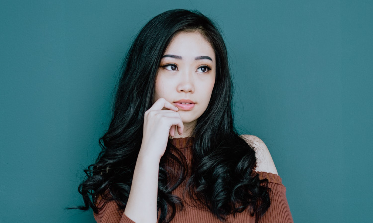 Young woman with dark silky hair stands against a teal wall wearing a rust-colored sweater with her hand ponderously touching her chin