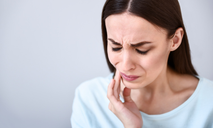 Closeup of a brunette woman in a blue shirt touching her cheek in pain due to an agonizing toothache