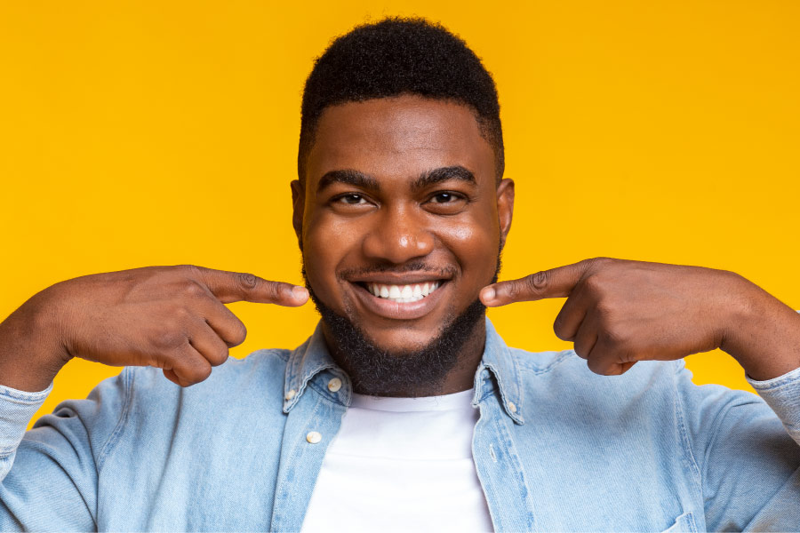 Dark-haired man smiles and points to his healthy gums against a yellow wall in Grafton, MA