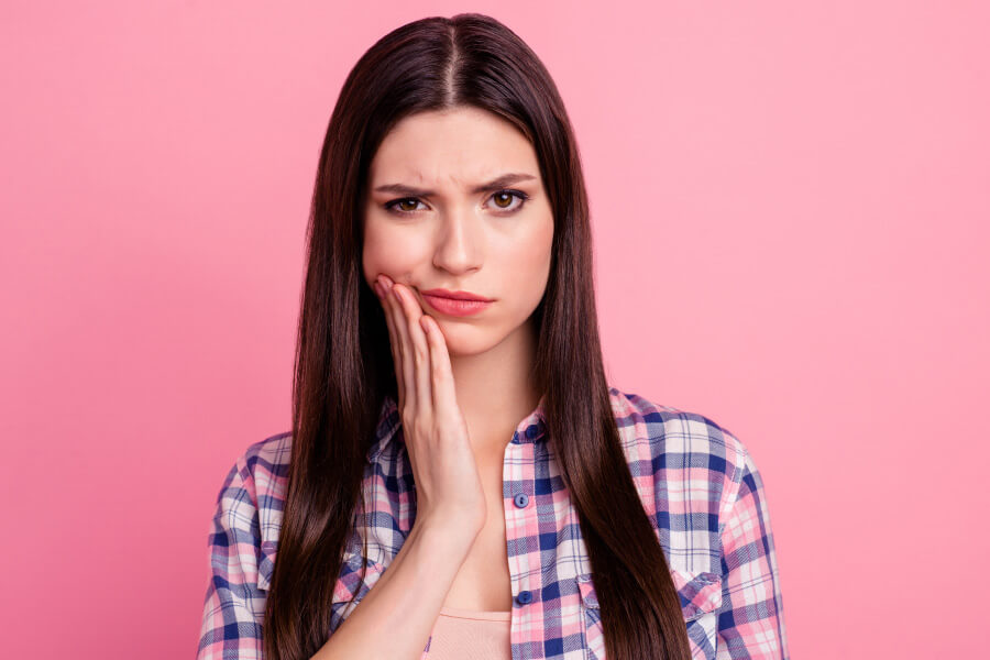 Brunette woman with a gumline cavity cringes in pain and touches her cheek against a pink wall