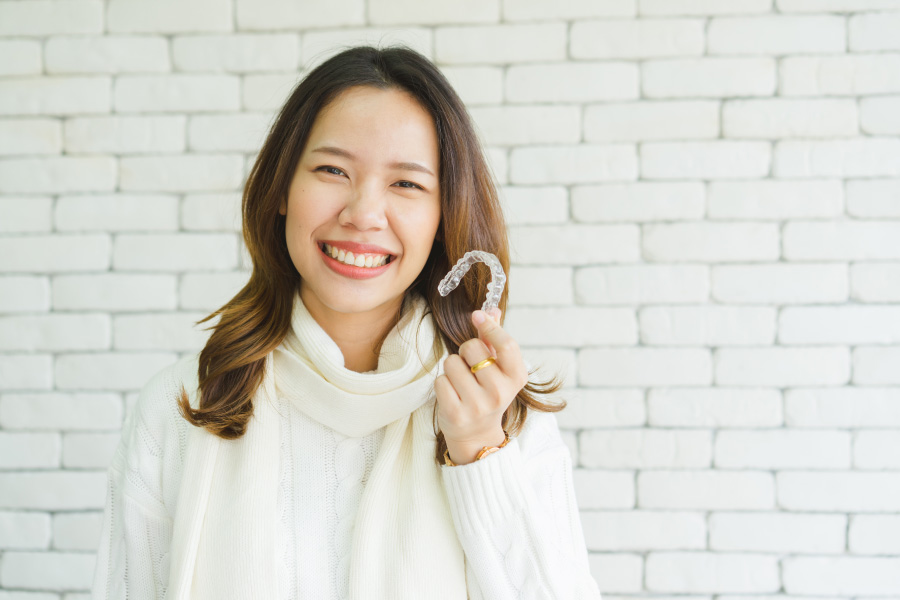 Brunette woman in a white sweater smiles while holding her Invisalign clear aligners to discuss their pros and cons