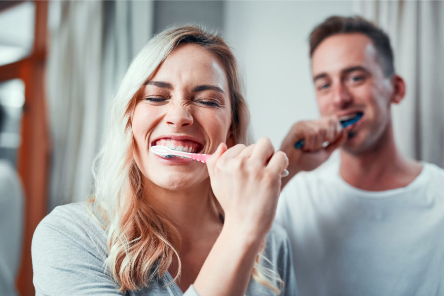 Closeup of a blonde woman brushing her teeth too hard next to her husband in the bathroom
