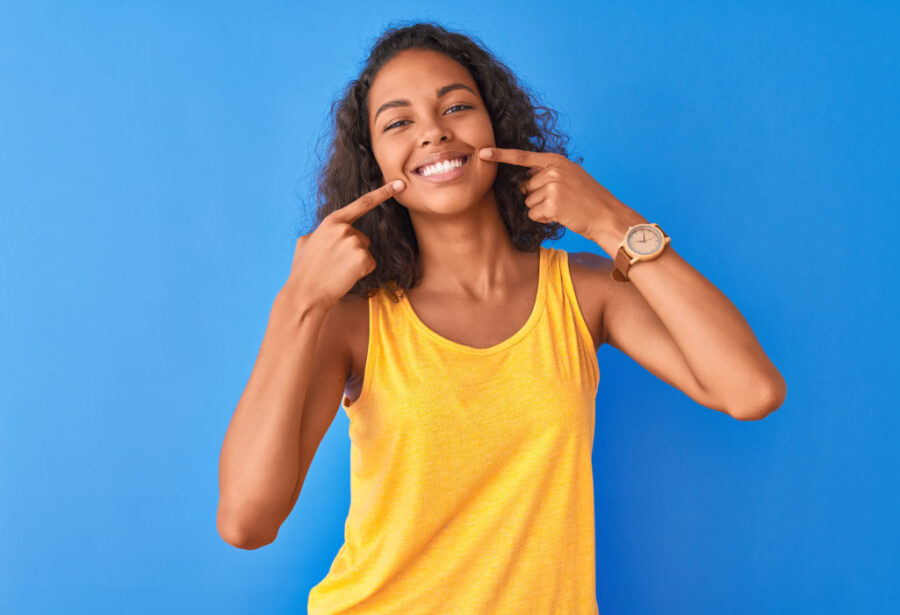 Curly-haired woman with veneers smiles and points to her teeth while wearing a yellow tank top against blue wall