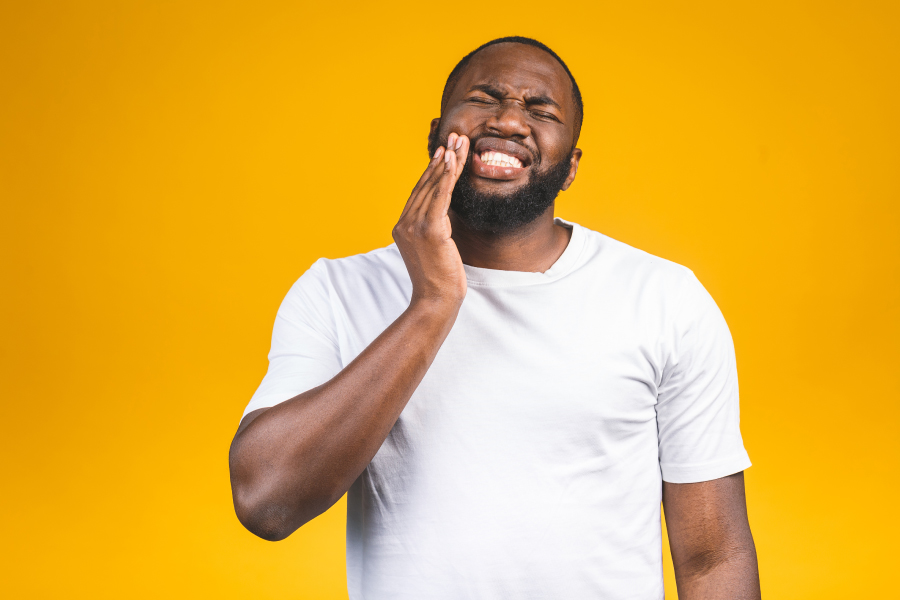 Black man in a white t-shirt cringes and touches his cheek due to severe tooth pain that needs an emergency dentist in Grafton, MA