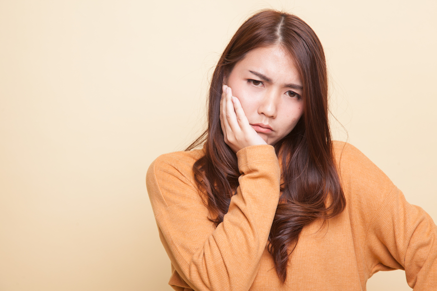 Brunette woman in a yellow sweater cringes in pain and holds her cheek due to a toothache